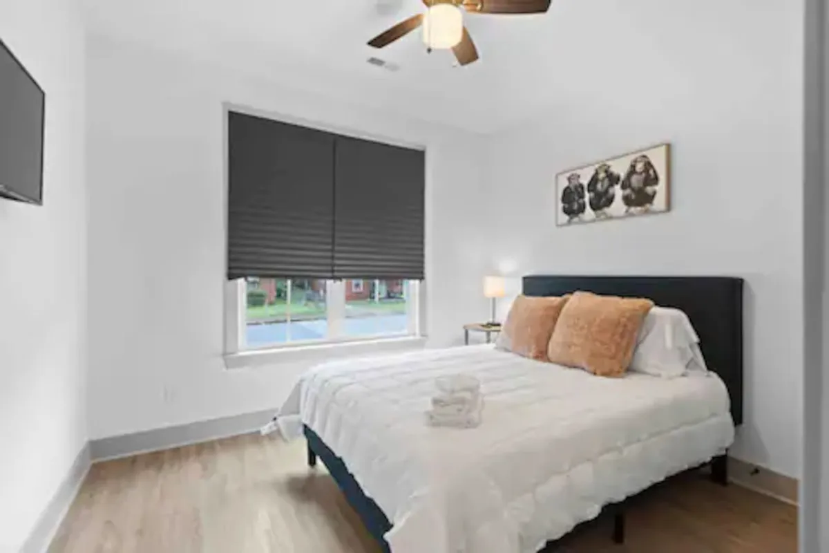 A plush bed with soft pillows sits beside a window with blinds, complemented by a bedside lamp and calming artwork. The room feels serene and private, inviting you to relax, read, or simply enjoy a quiet moment.