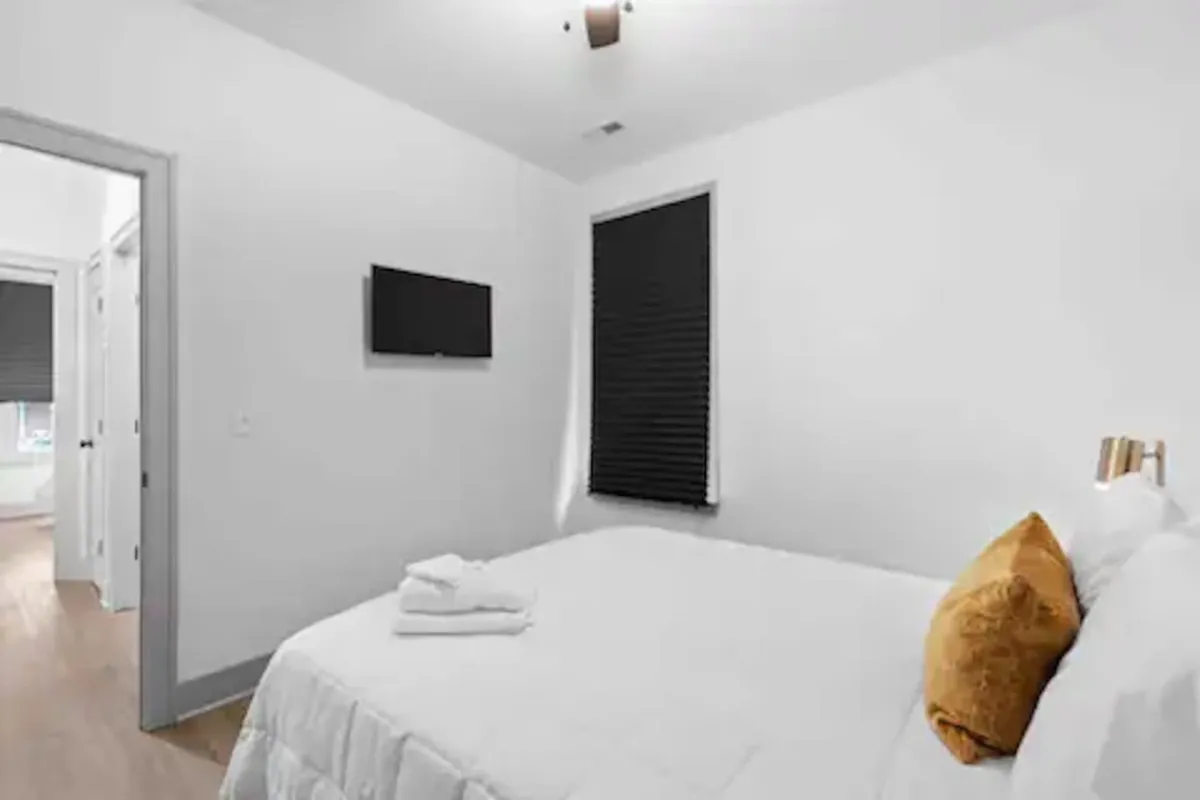 A serene bedroom with a bed dressed in crisp white linens, a gold accent pillow, and a wall-mounted TV. The room is softly lit, conveying a sense of calm and restfulness.