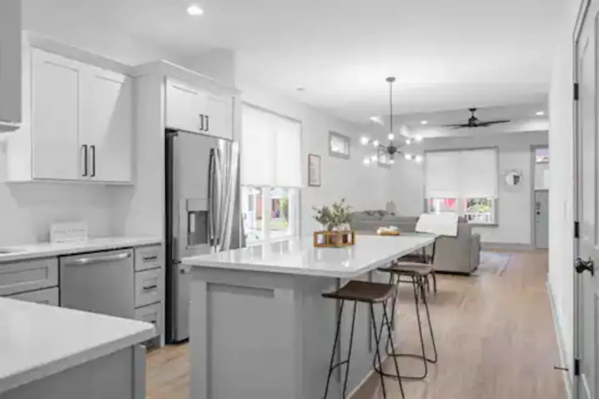 A modern, open living area featuring a kitchen island with stylish bar stools and stainless steel appliances. The view extends into a comfortable living space in the background, emphasizing the open-concept design and seamless flow between the gathering areas.