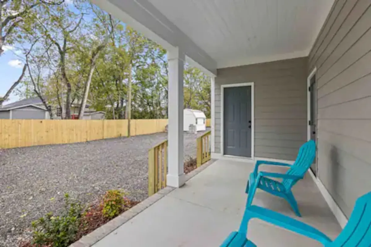 The image presents the exterior of the property, featuring blue porch chairs, a front door, a gravel driveway, and a fenced yard. The setting captures a welcoming, peaceful mood, with morning sunlight filtering through the trees and a spacious, inviting porch.