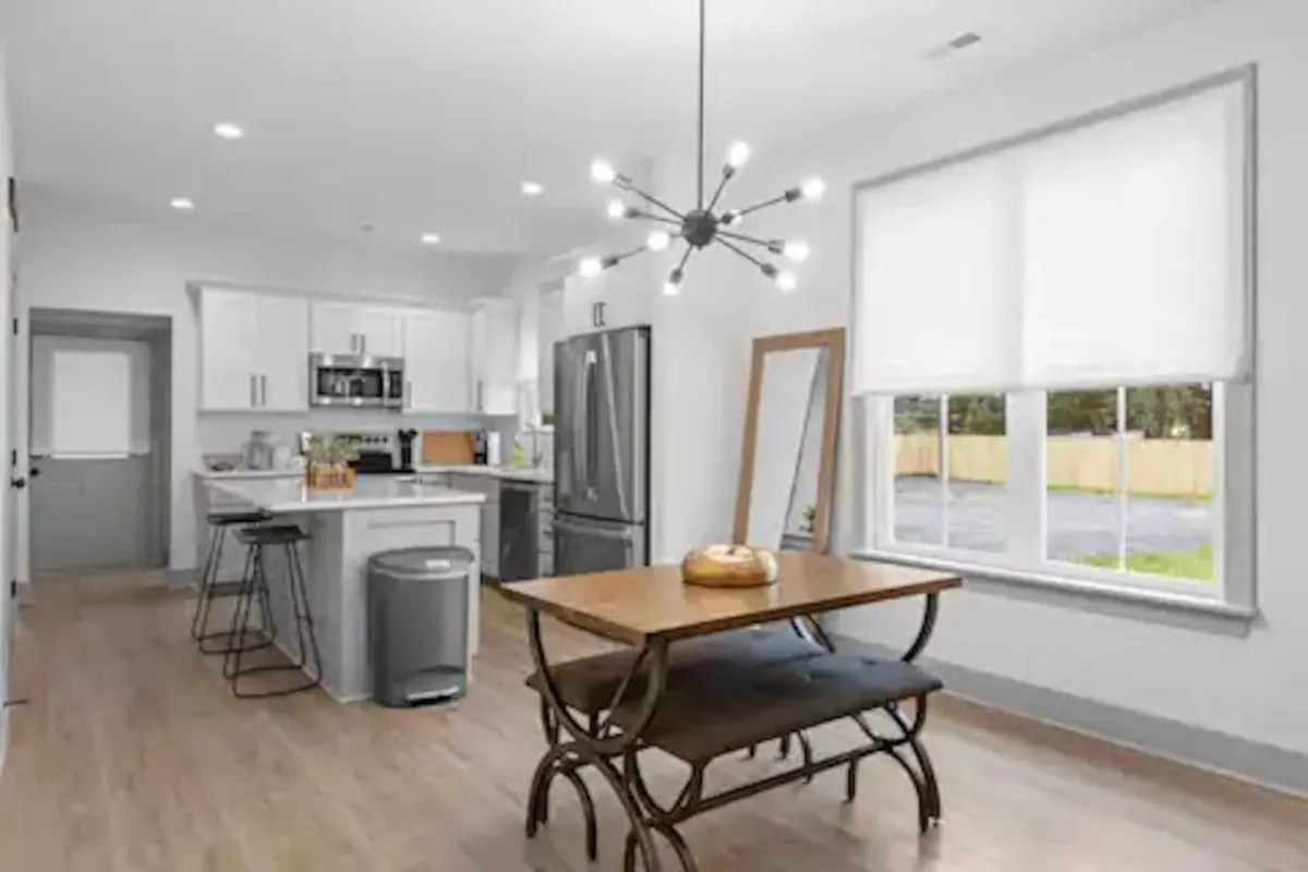 A stylish dining area with a modern light fixture, a large dining table, and a kitchen island with appliances. Sunlight streams through a window, illuminating the space and contributing to a sense of openness and energy efficiency.