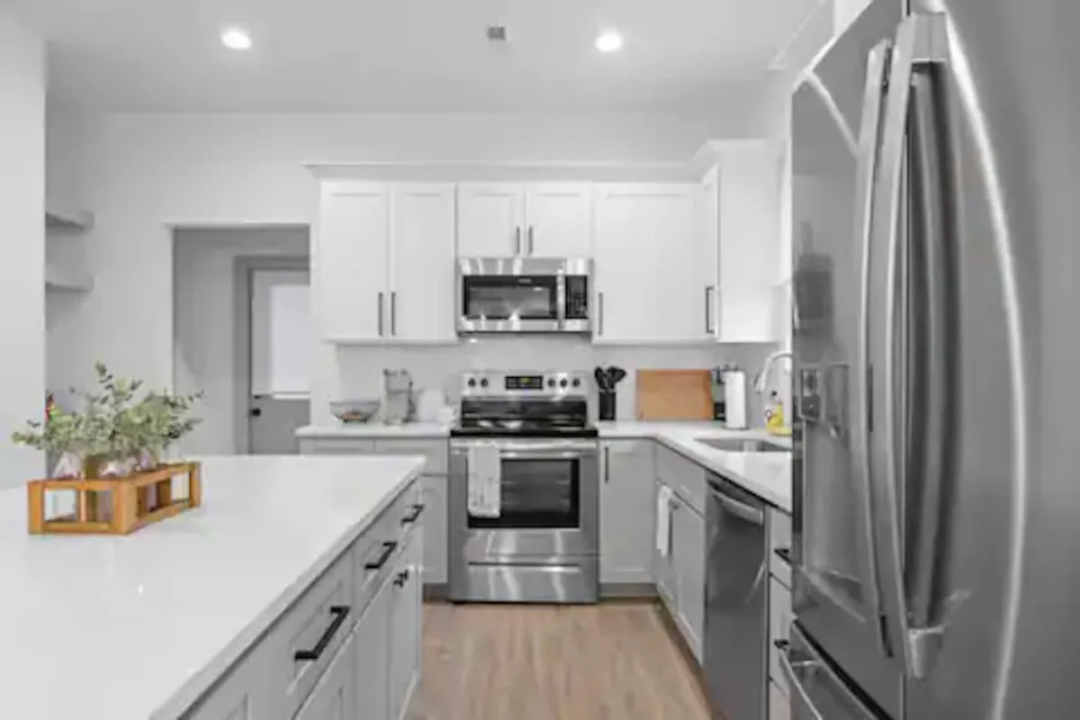 A bright, modern kitchen with stainless steel appliances, white cabinetry, and a central kitchen island. The space is arranged for both cooking and gathering, with plenty of counter space and a warm, welcoming atmosphere.