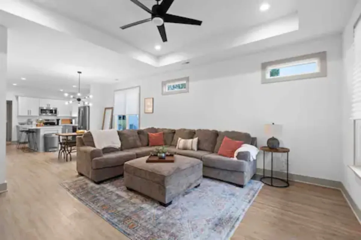 A spacious living room bathed in natural light, centered around a large sectional sofa, decorative ottoman, and a colorful area rug. A window lets in sunlight, creating a bright and inviting atmosphere ideal for relaxation and socializing.